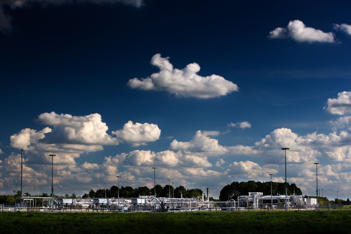 Een bewolkte blauwe lucht boven een grasveld met elektriciteitsleidingen en kleine gebouwen aan de horizon.