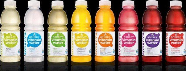 Acht kleurrijke flessen drank op een rij tegen een zwarte achtergrond.