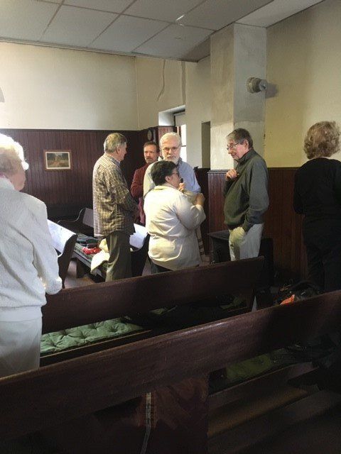 People conversing inside a room, possibly a church. Wooden benches in the foreground, dark paneling on wall.