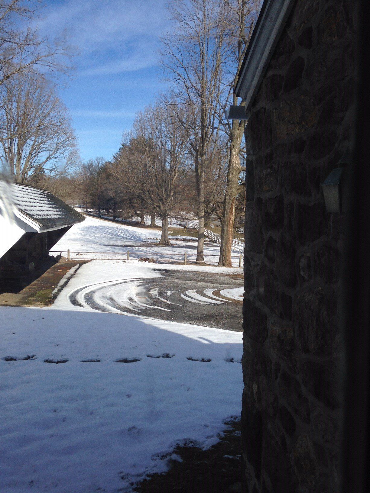 Snowy driveway next to a stone building with bare trees under a blue sky.