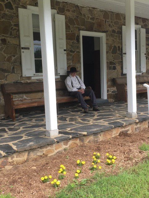 Man in old-fashioned clothes sits on a bench on the porch of a stone building, yellow flowers in front.