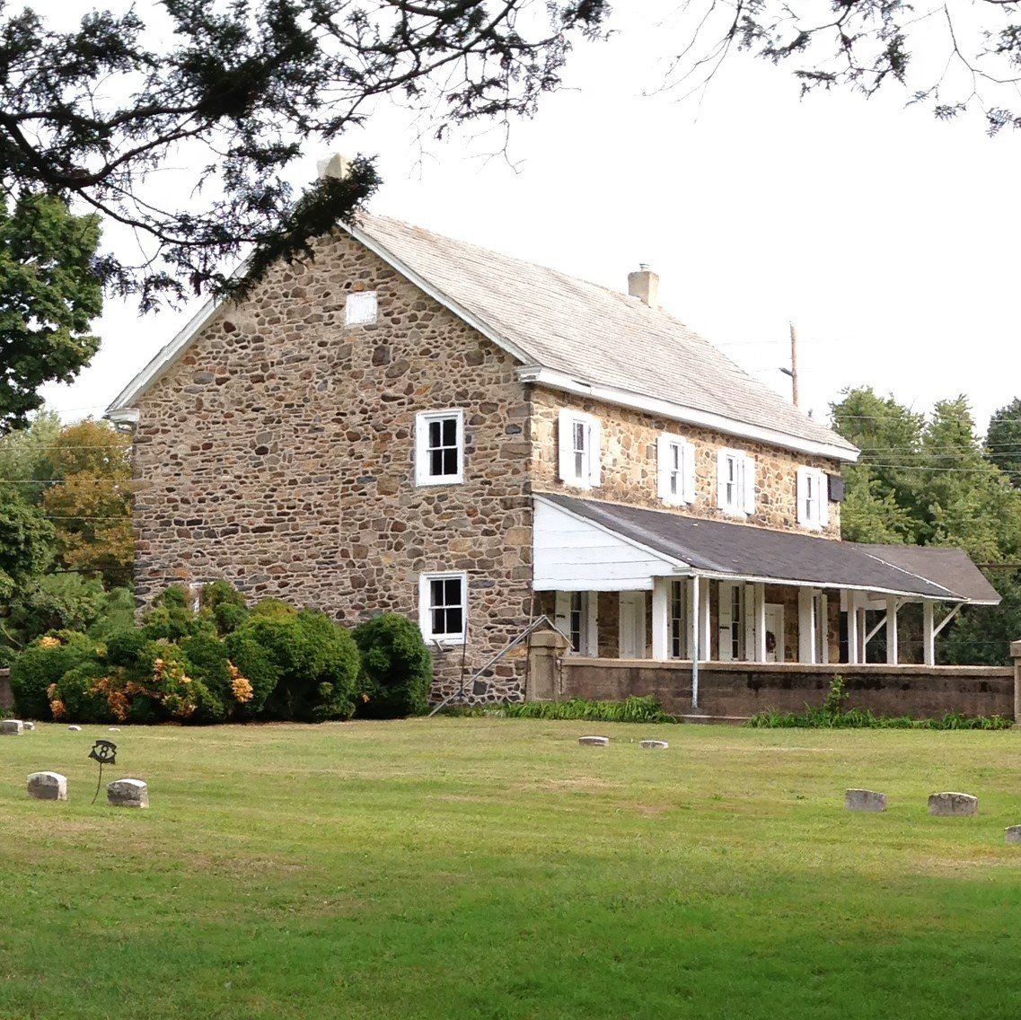 Stone building with porch and white shutters, next to a grassy lawn with tombstones.