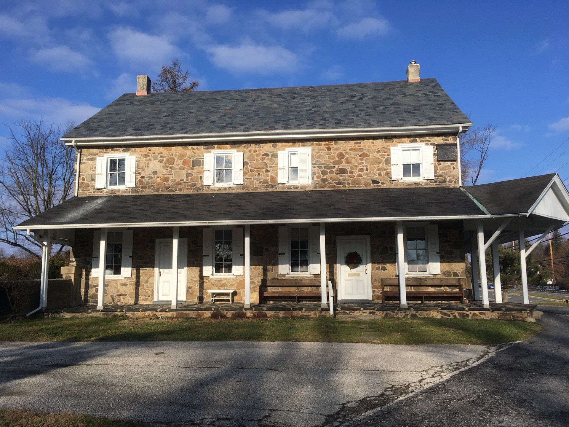 Stone farmhouse with porch, white shutters, and a gray roof under a blue sky.