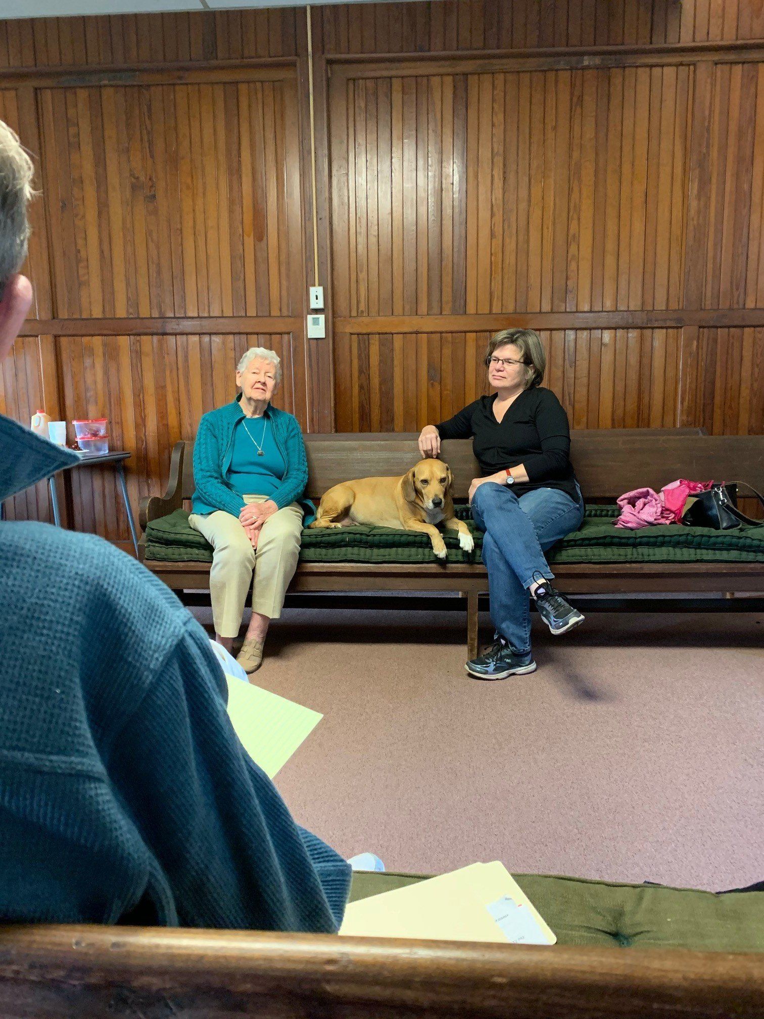 Two women with a dog on a church pew; one petting the dog. A person in foreground. Dark wood paneling.