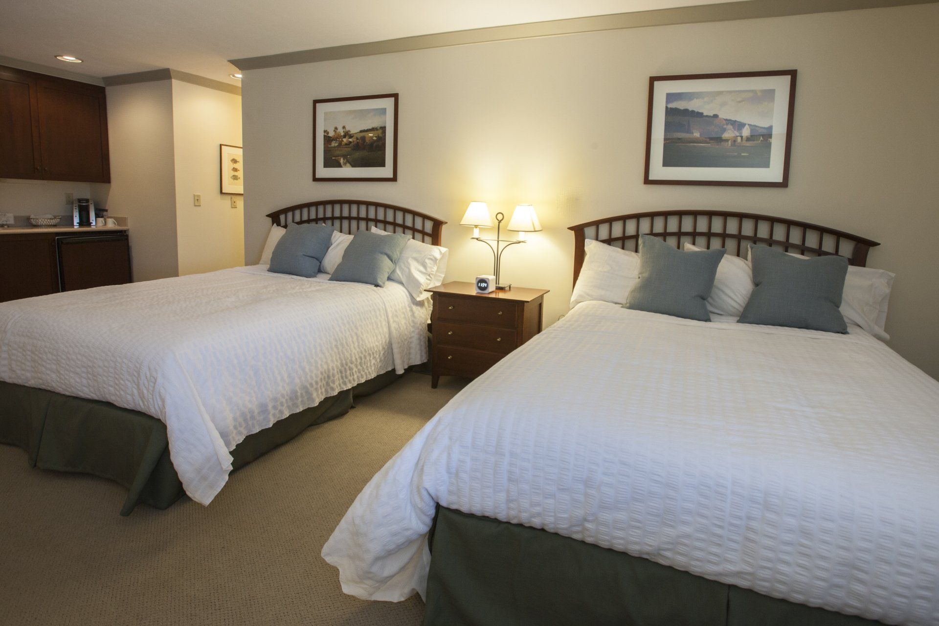 Two queen size beds with a night stand in between the beds with a lamp and an alarm clock on it.