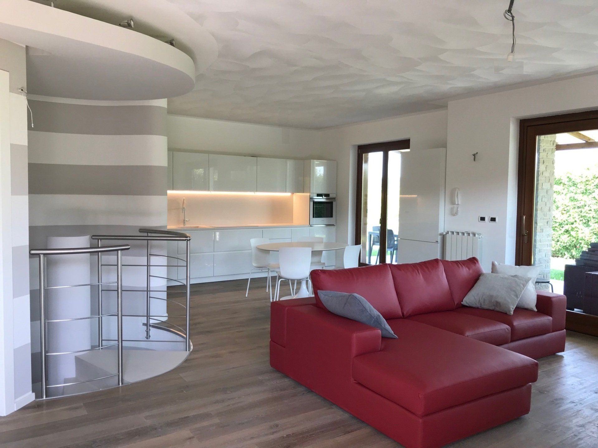 Moderno spazio abitativo open space con divano rosso, cucina bianca e scala a chiocciola con ringhiere in metallo.