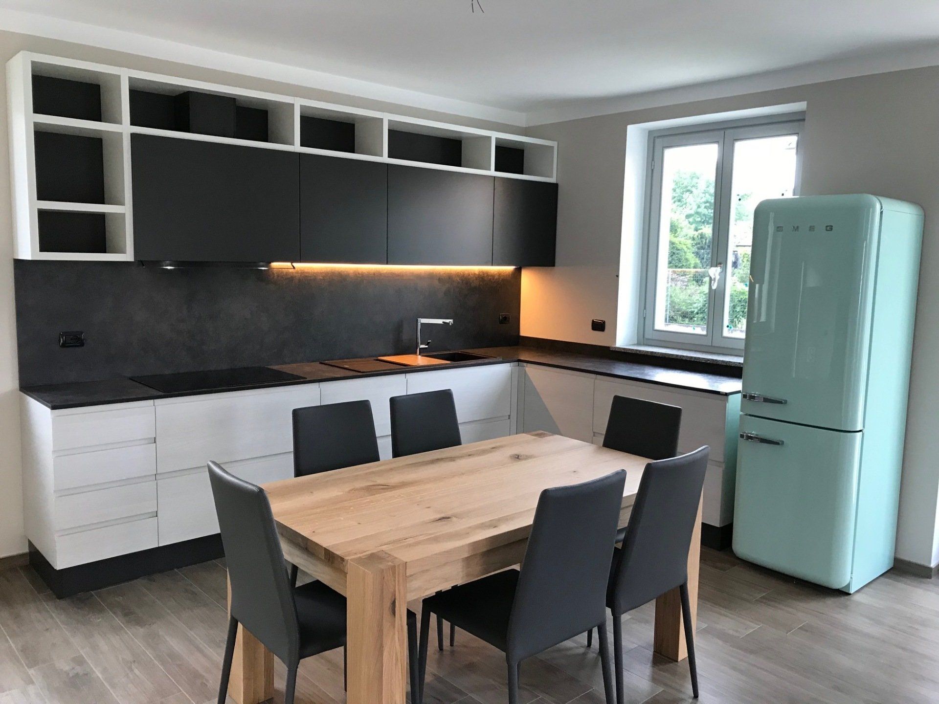 Cucina moderna con mobili bianchi e neri, tavolo in legno, sedie grigie e frigorifero verde menta.