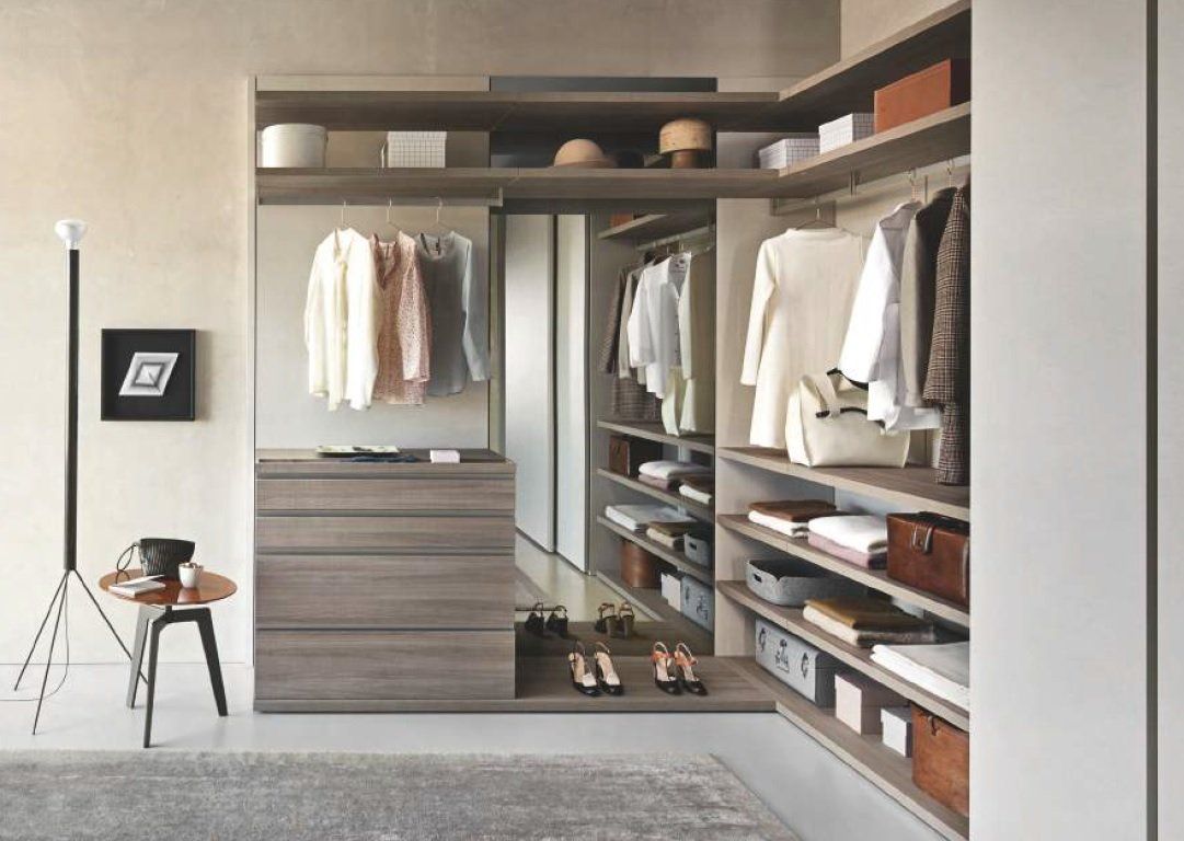 Cabina armadio con ripiani organizzati, abiti appesi e cassetti. Colori neutri e una piccola sedia con treppiede.