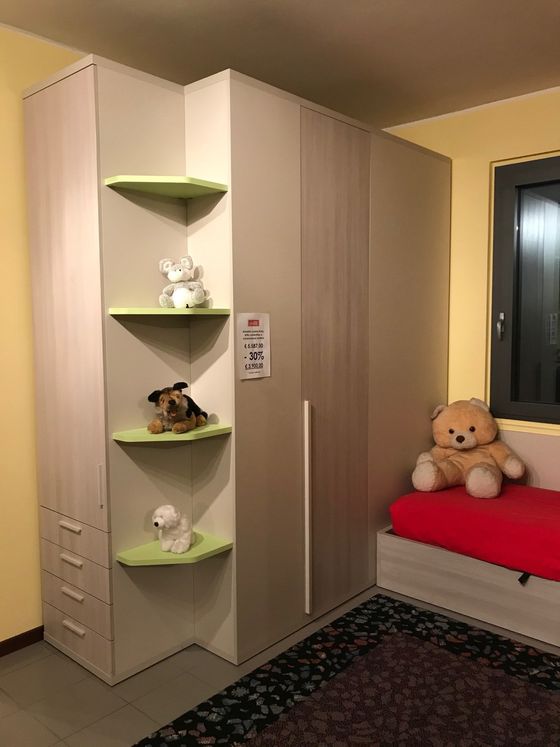 Armadio angolare con ripiani, beige e verde, animali di peluche esposti, accanto a un letto e una finestra.