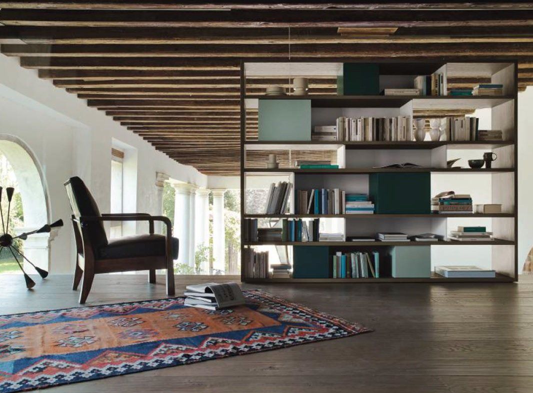 Soggiorno con libreria, tappeto, sedia e soffitto in legno a vista.