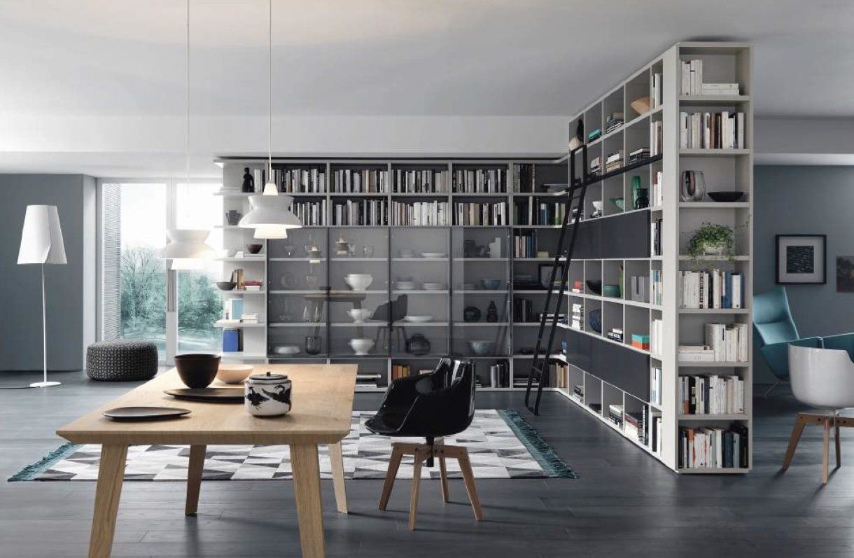 Soggiorno moderno con librerie integrate che dividono lo spazio. Tavolo, sedie e scala in legno chiaro. Pareti grigie.
