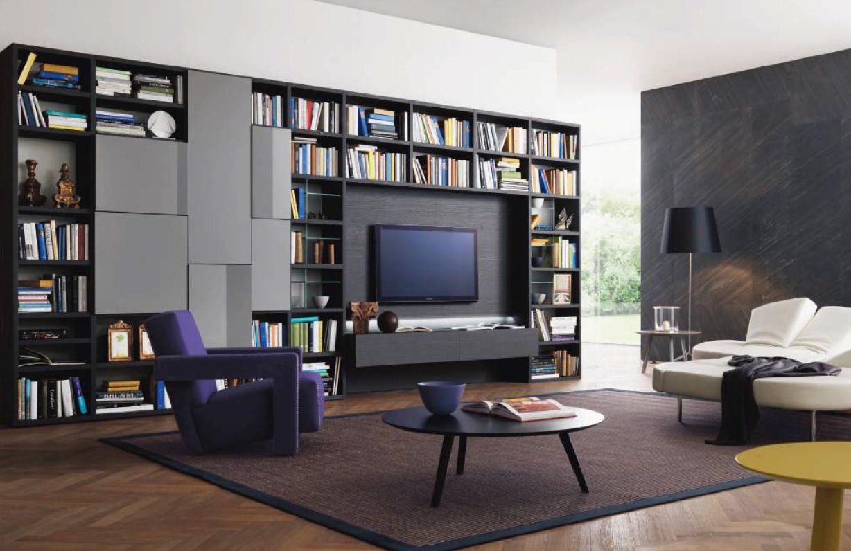 Soggiorno con libreria scura integrata attorno alla TV, sedia viola, mobili moderni e tappeto a fantasia.