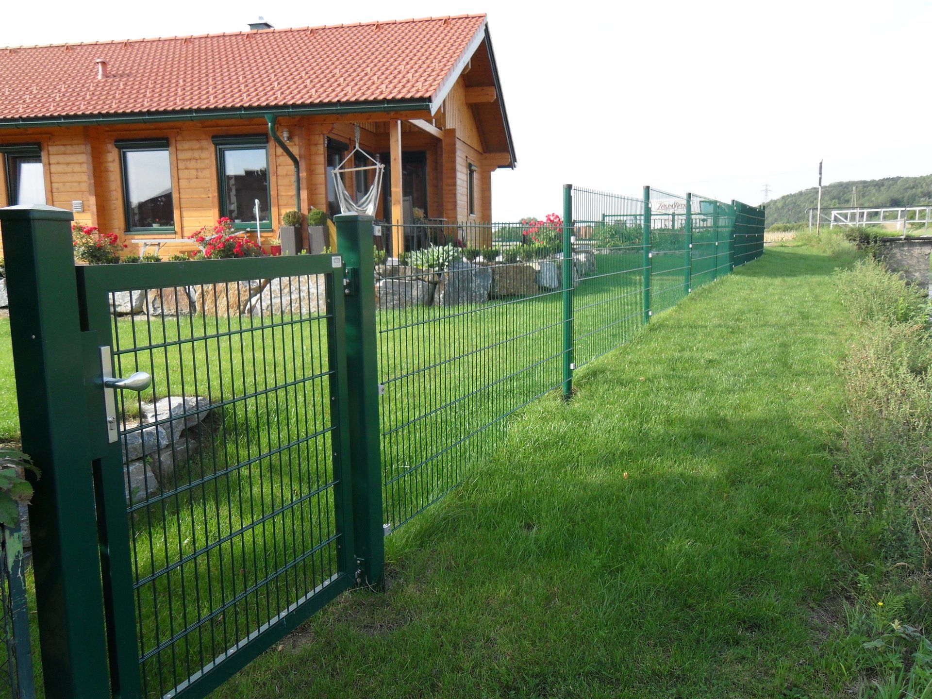 Ein grüner Zaun umgibt einen üppig grünen Hof vor einem Holzhaus.