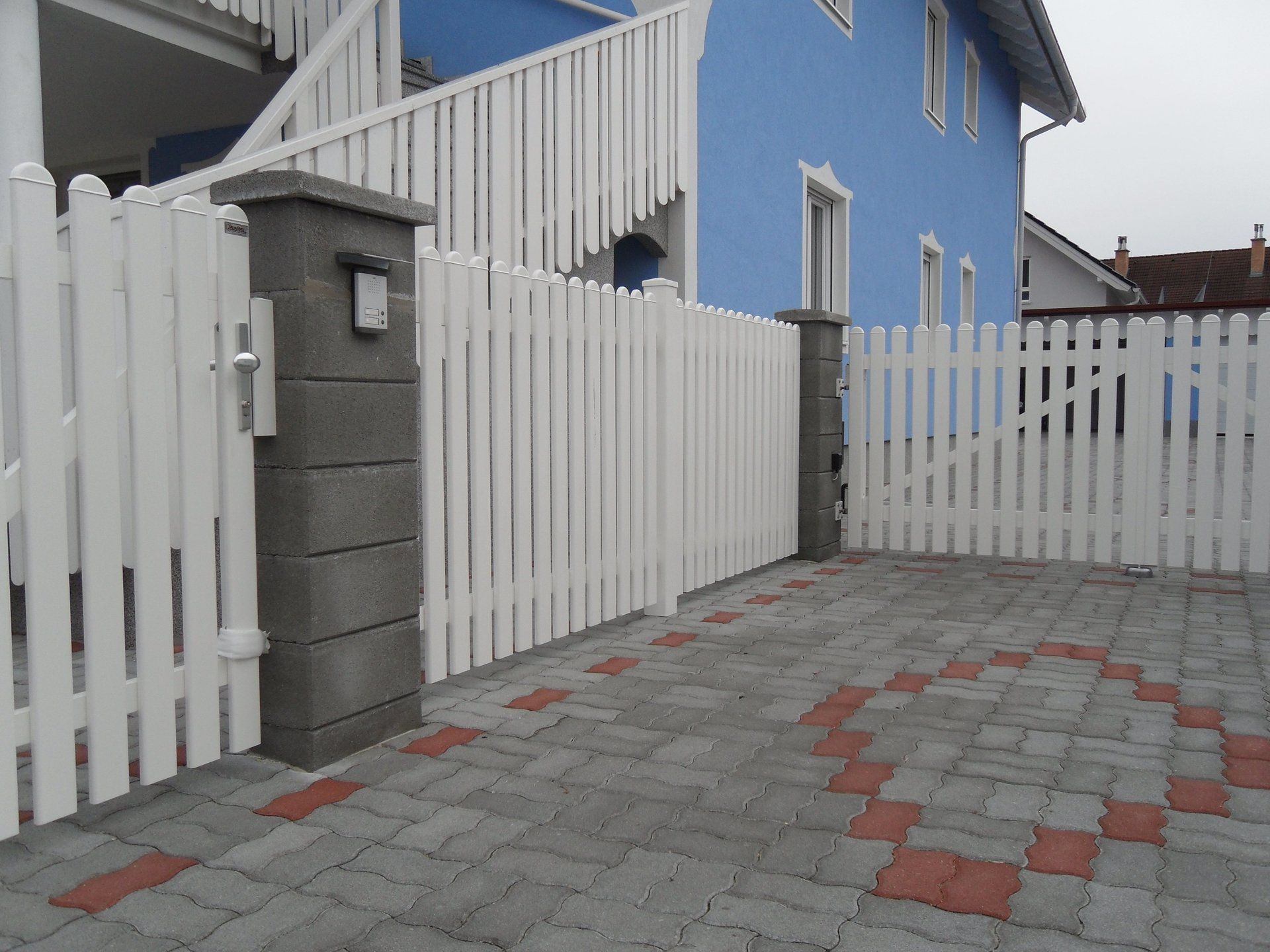 Ein weißer Lattenzaun umgibt eine gemauerte Auffahrt vor einem blauen Haus