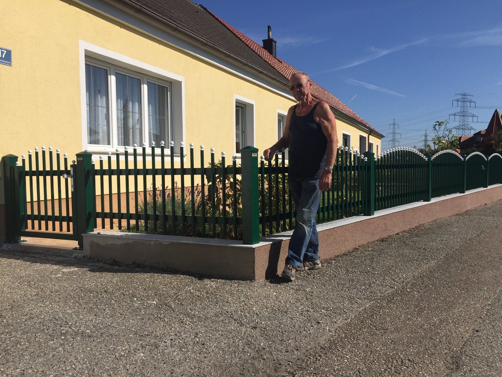Ein Mann steht neben einem grünen Zaun vor einem gelben Haus.