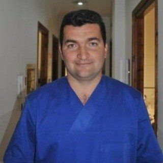 Uomo in camice blu, sorridente in un ambiente interno, probabilmente un ambiente sanitario.