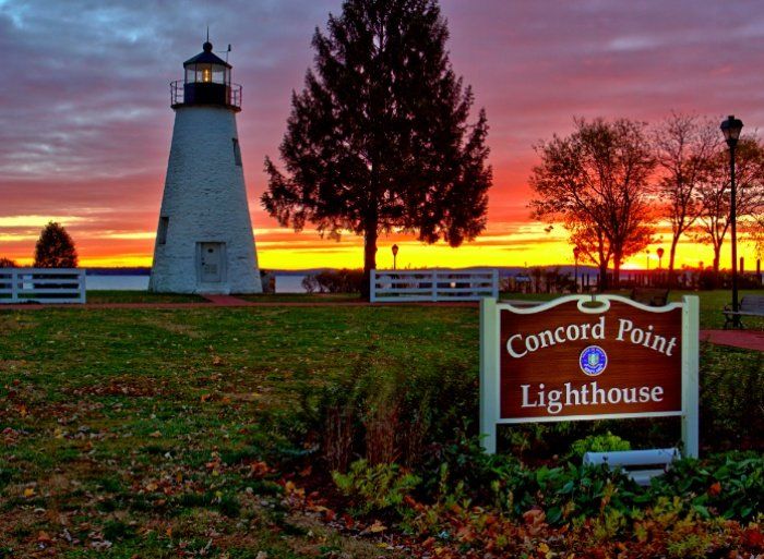 Concord Point Lighthouse at sunset; white tower, orange sky, green grass, sign.