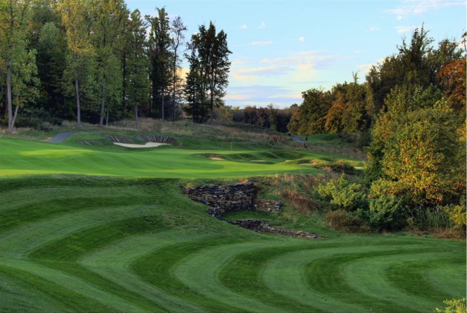 Green golf course with curved mowing patterns, trees, and a bunker.