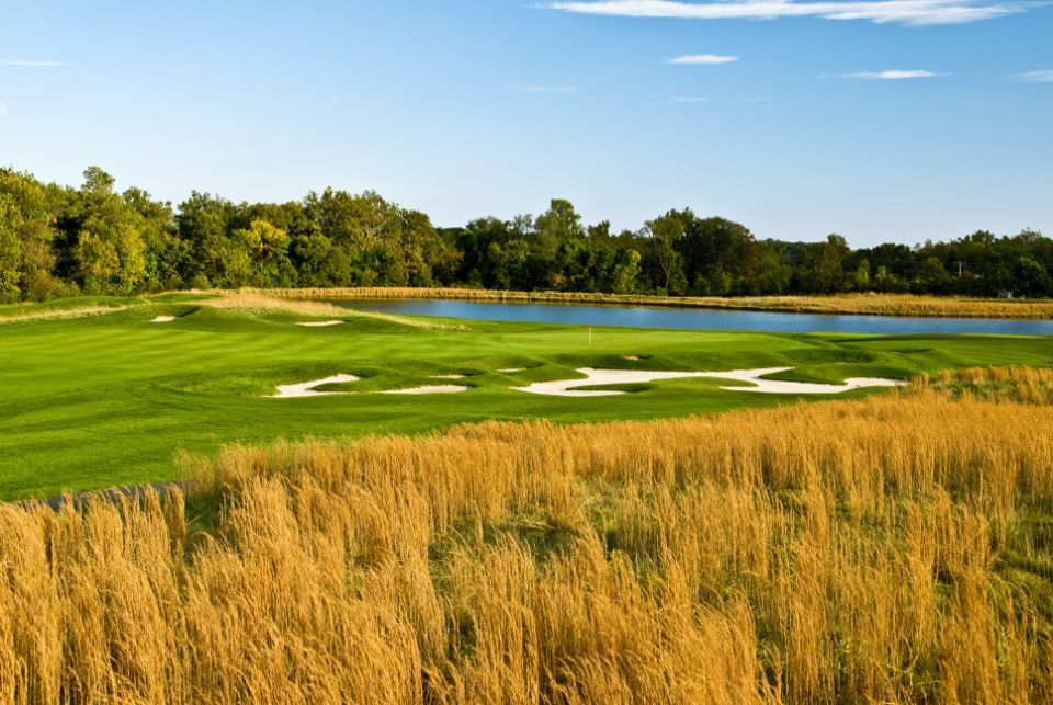 Golf course scene with green grass, water, and tall yellow grass in foreground. Blue sky and trees in background.