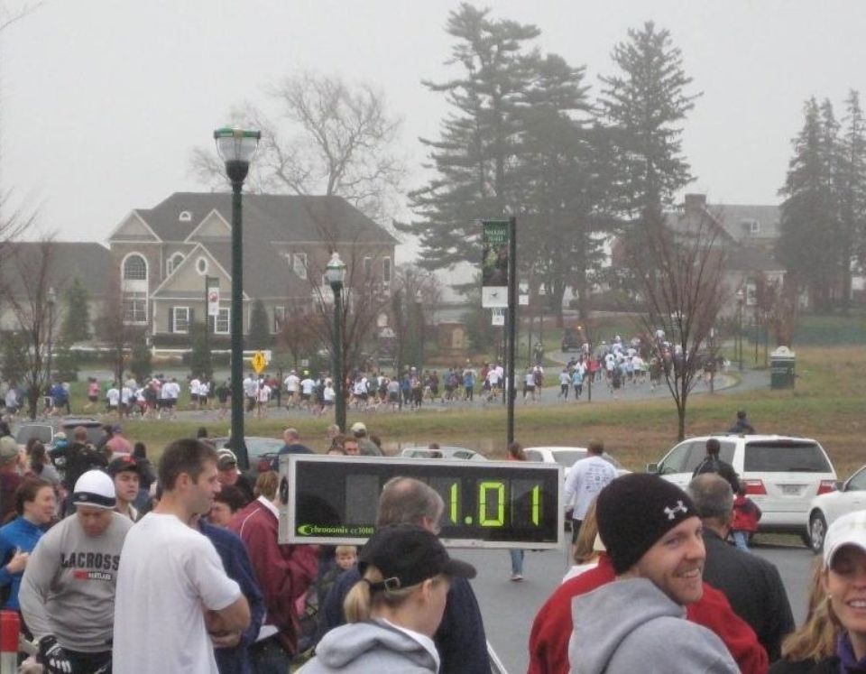 Runners in a race, a digital clock displays 1.01 mile mark, crowd watches on a cloudy day.