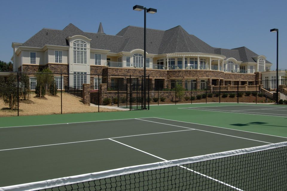 Tennis court in foreground; large, multi-story building in background. Dark green court with net. Blue sky.