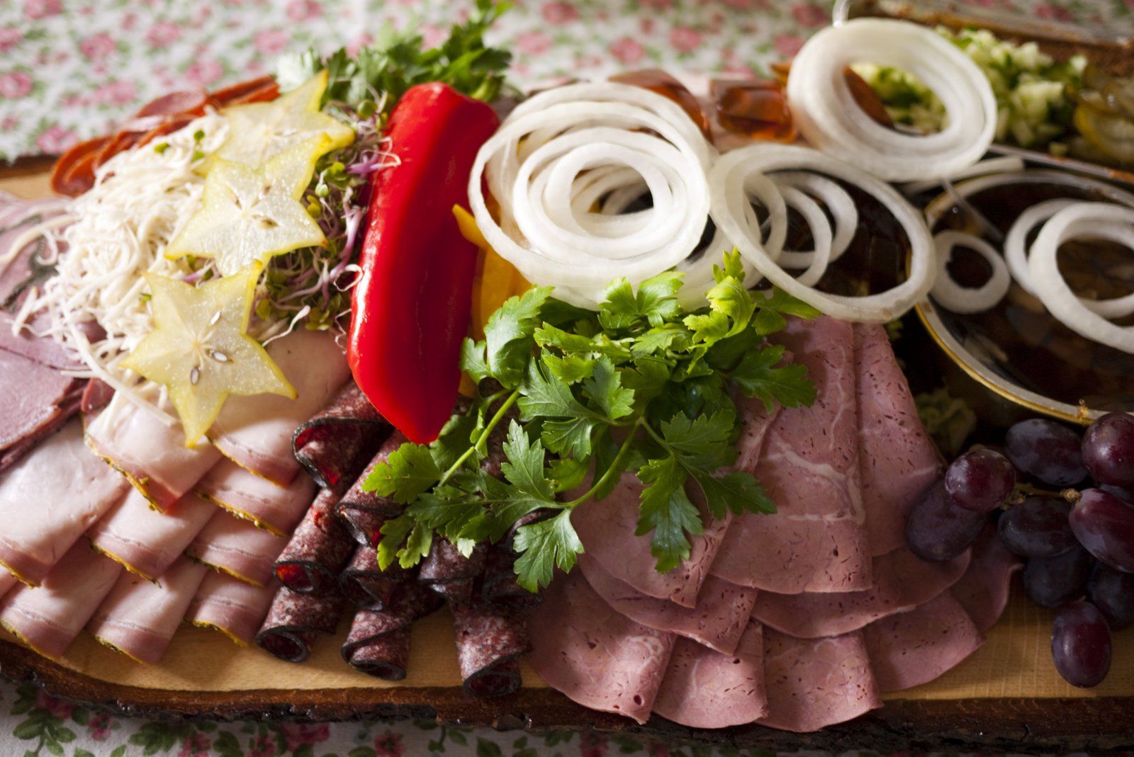 A wooden cutting board topped with meat , onions , grapes and peppers.