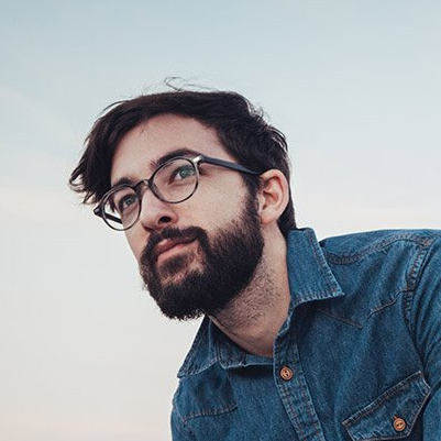 Man with glasses and a beard in a denim shirt looks upwards, with a pale blue sky background.