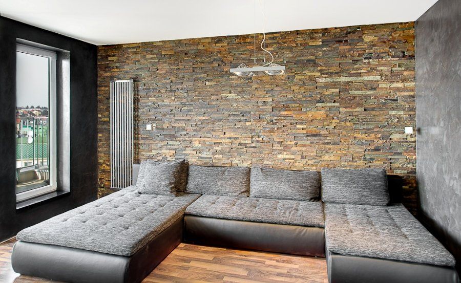 Nowoczesny salon z szarą, narożną sofą na tle kamiennej ściany i czarnymi ścianami bocznymi.