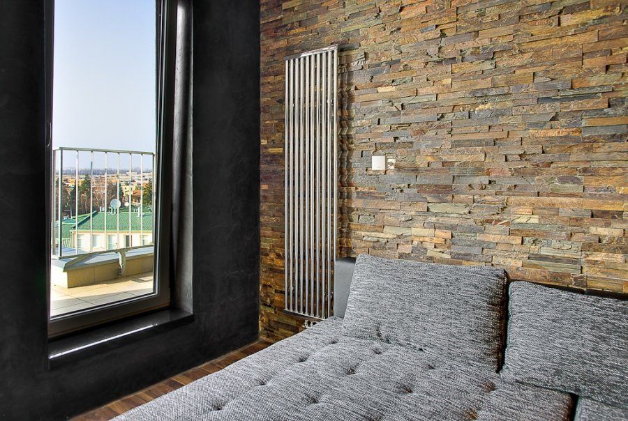 Pokój z kamienną ścianą, oknem, szarą kanapą i wysokim srebrnym kaloryferem. Widok na balkon.