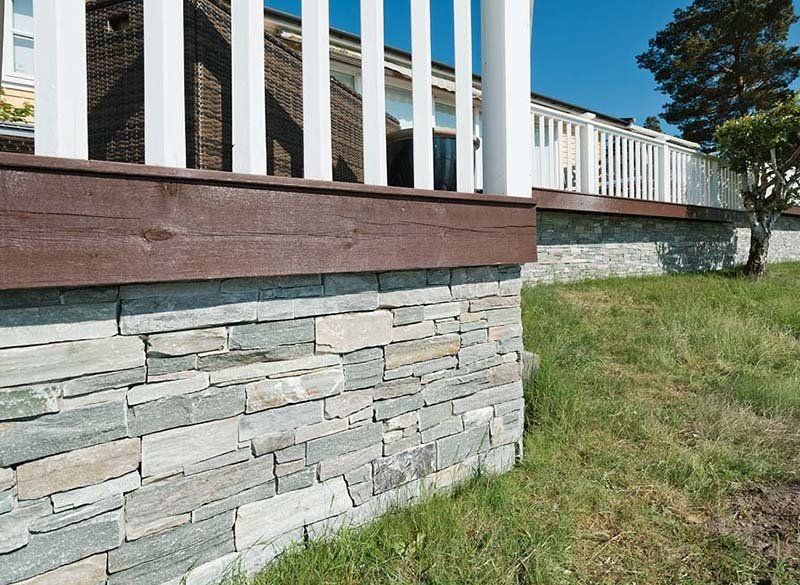 Kamienny mur oporowy podtrzymuje taras z białymi balustradami i brązowymi drewnianymi wykończeniami. Na pierwszym planie znajduje się zielona trawa.