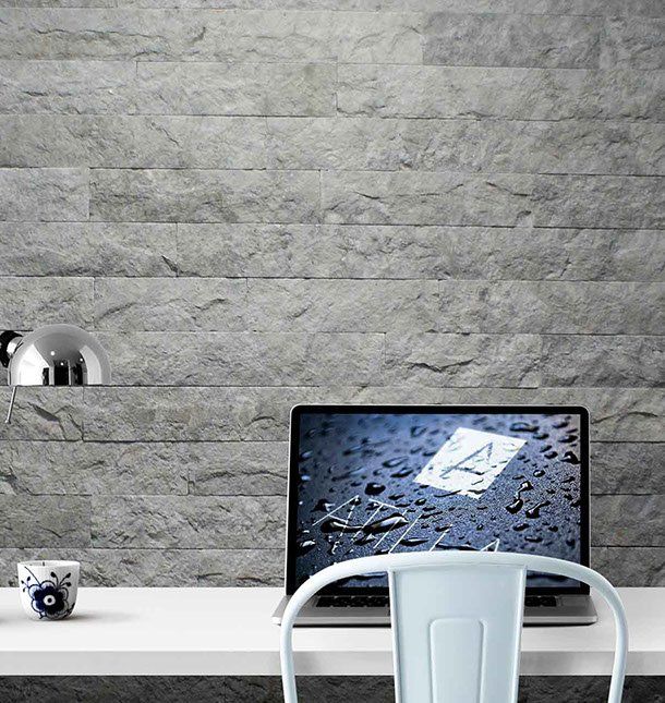 Laptop na białym biurku na tle szarej kamiennej ściany; niebieskie krzesło i kubek.