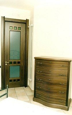 Porta e cassettiera in legno scuro in una stanza con pareti bianche, pavimento in piastrelle color cuoio e un termosifone in metallo.