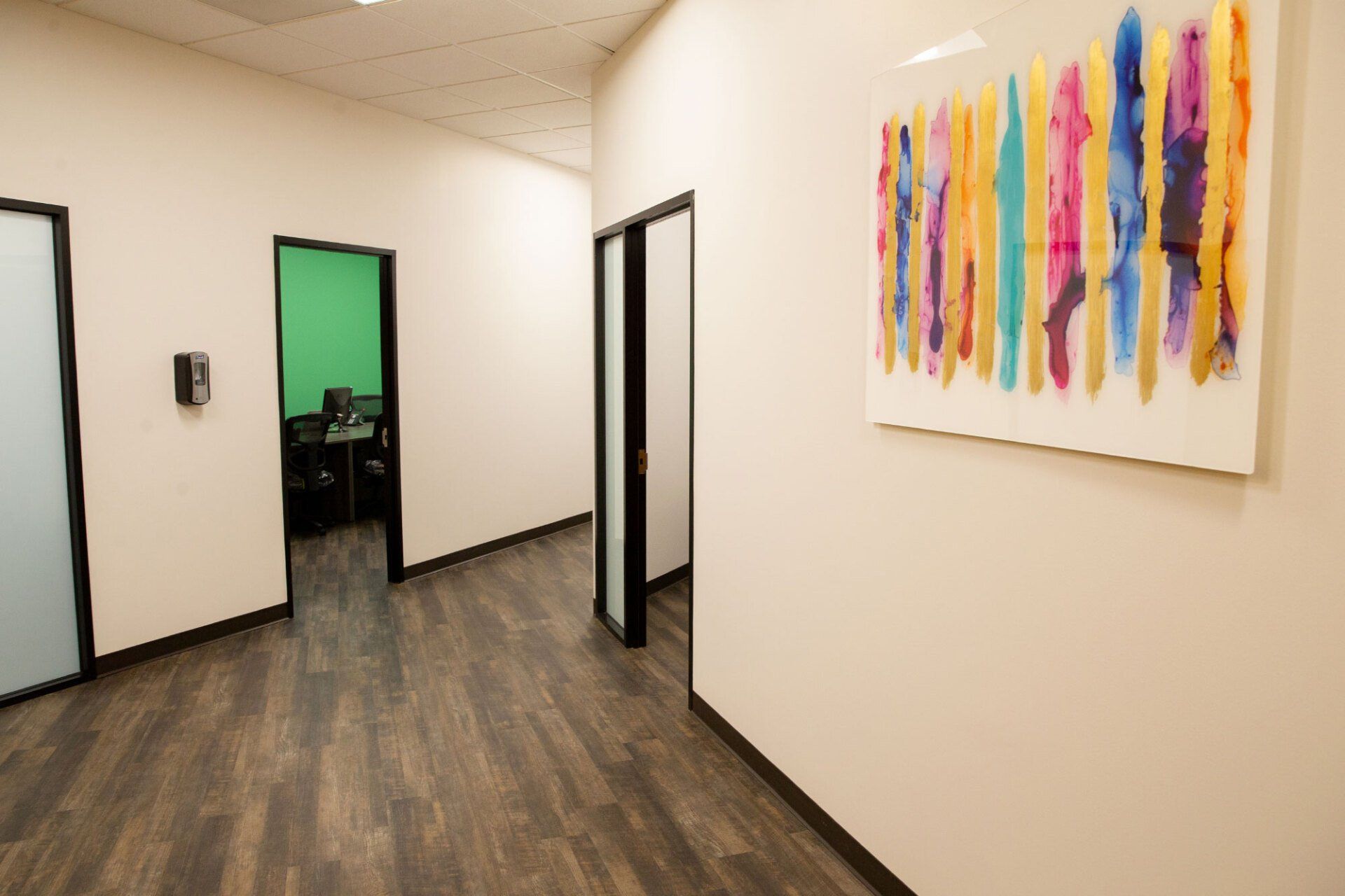 A hallway with light walls and wood-patterned flooring, featuring a colorful abstract art piece and several open doors.