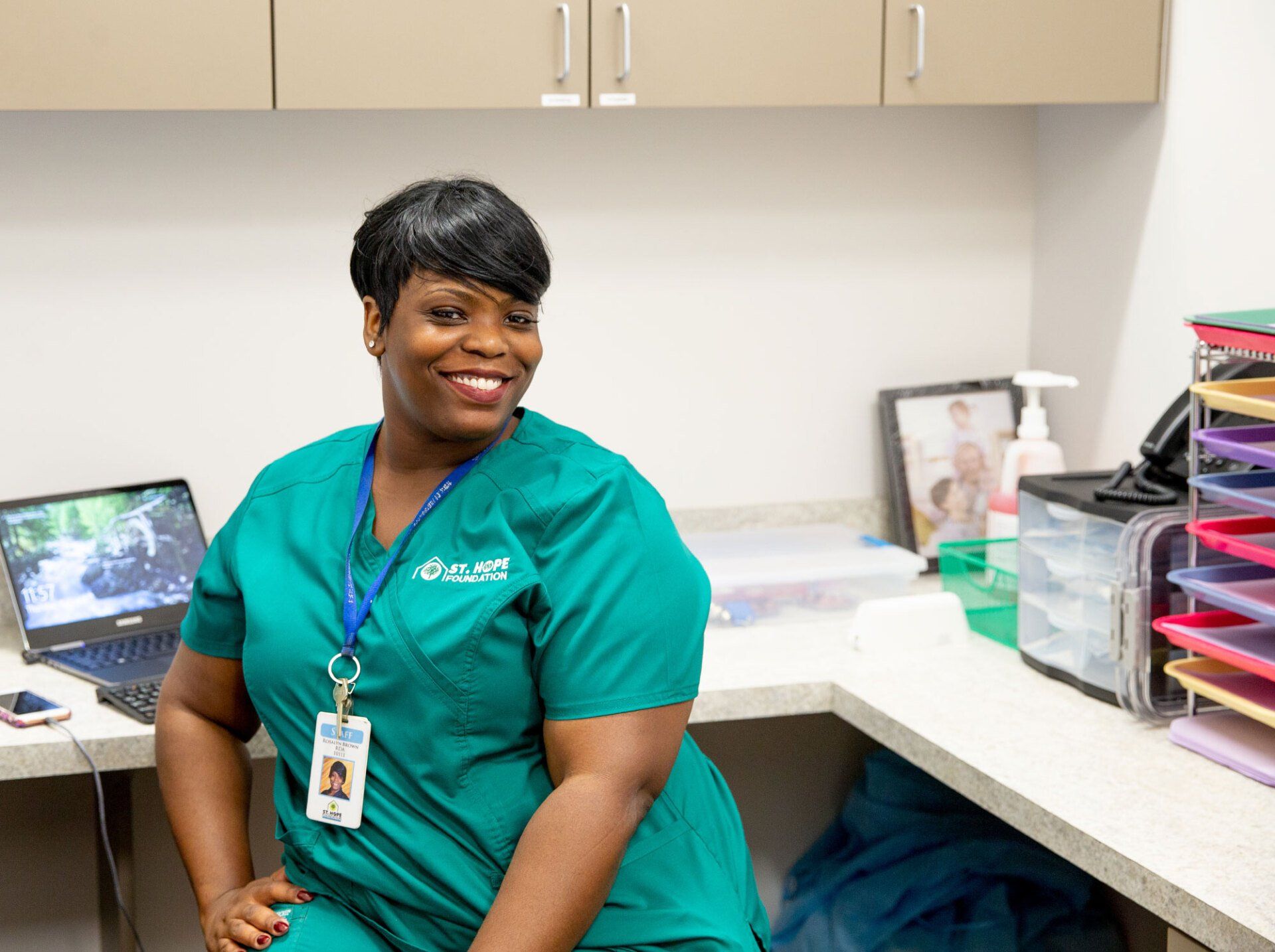 A smiling professional in green medical scrubs sits at a desk in an office with a laptop and paper tray.