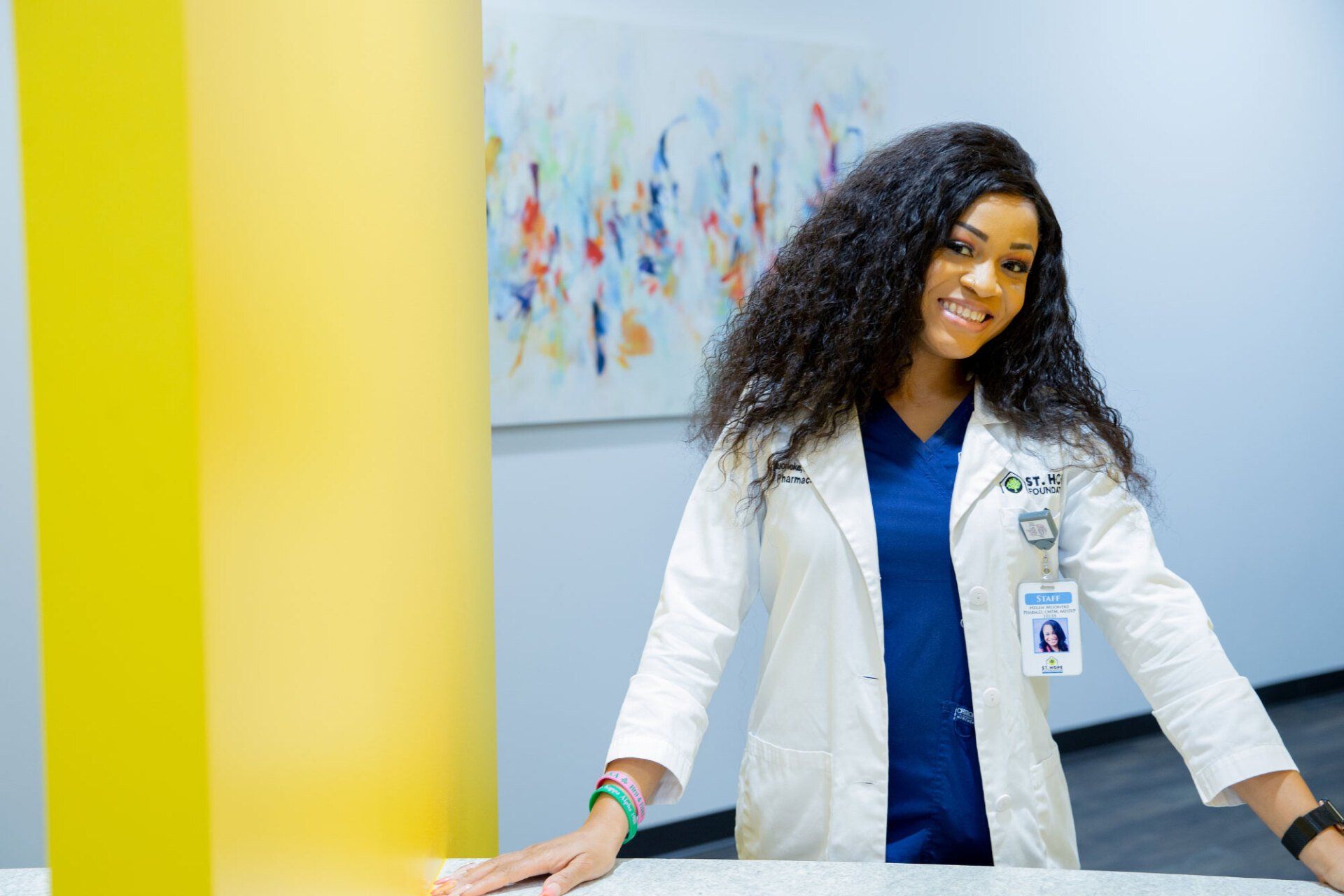 A smiling healthcare worker in a white coat and blue scrubs leans against a counter next to a bright yellow pillar.