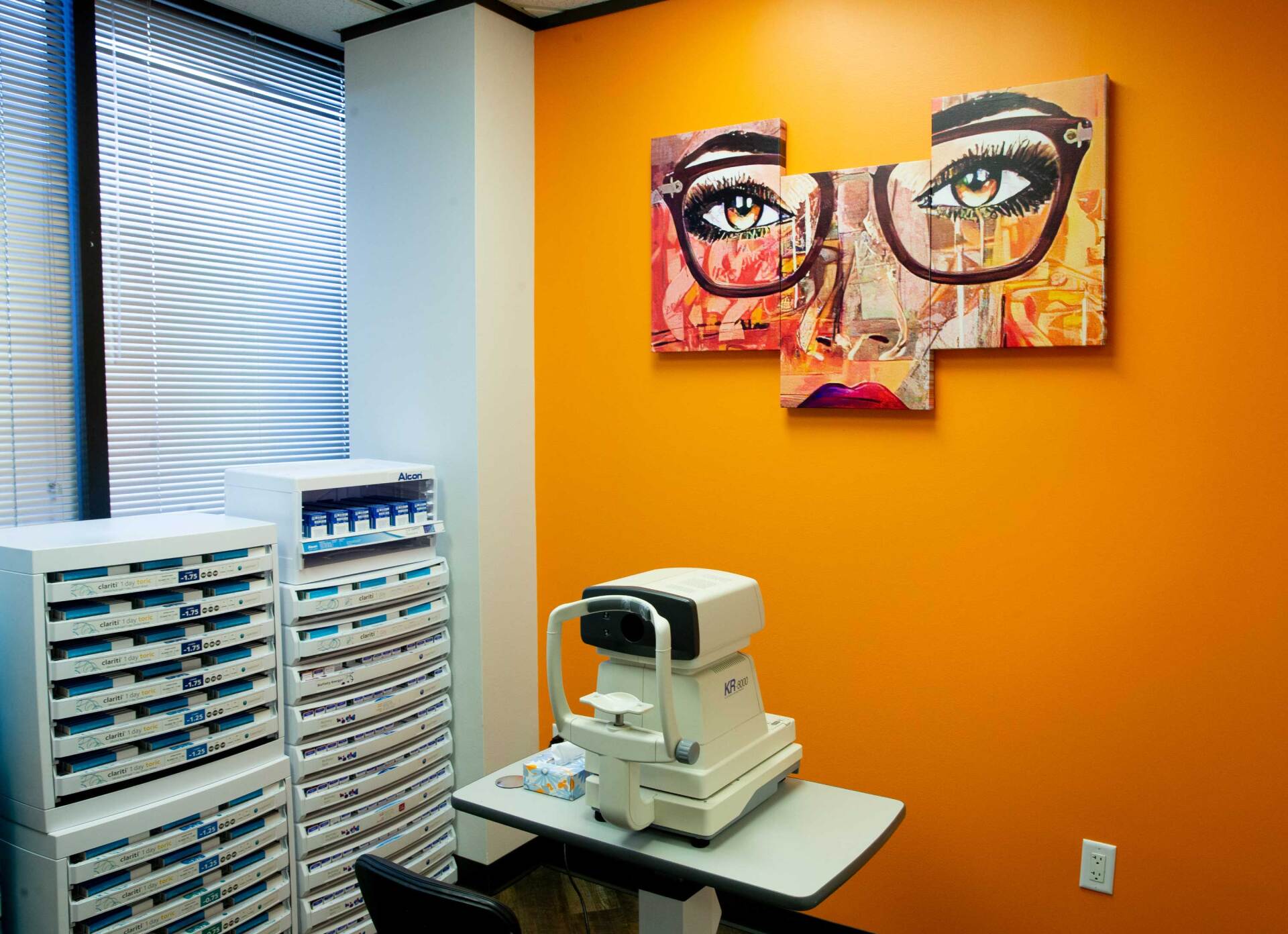 An optometrist's exam room with an orange wall, a white eye-exam machine, and abstract art of eyes wearing glasses.