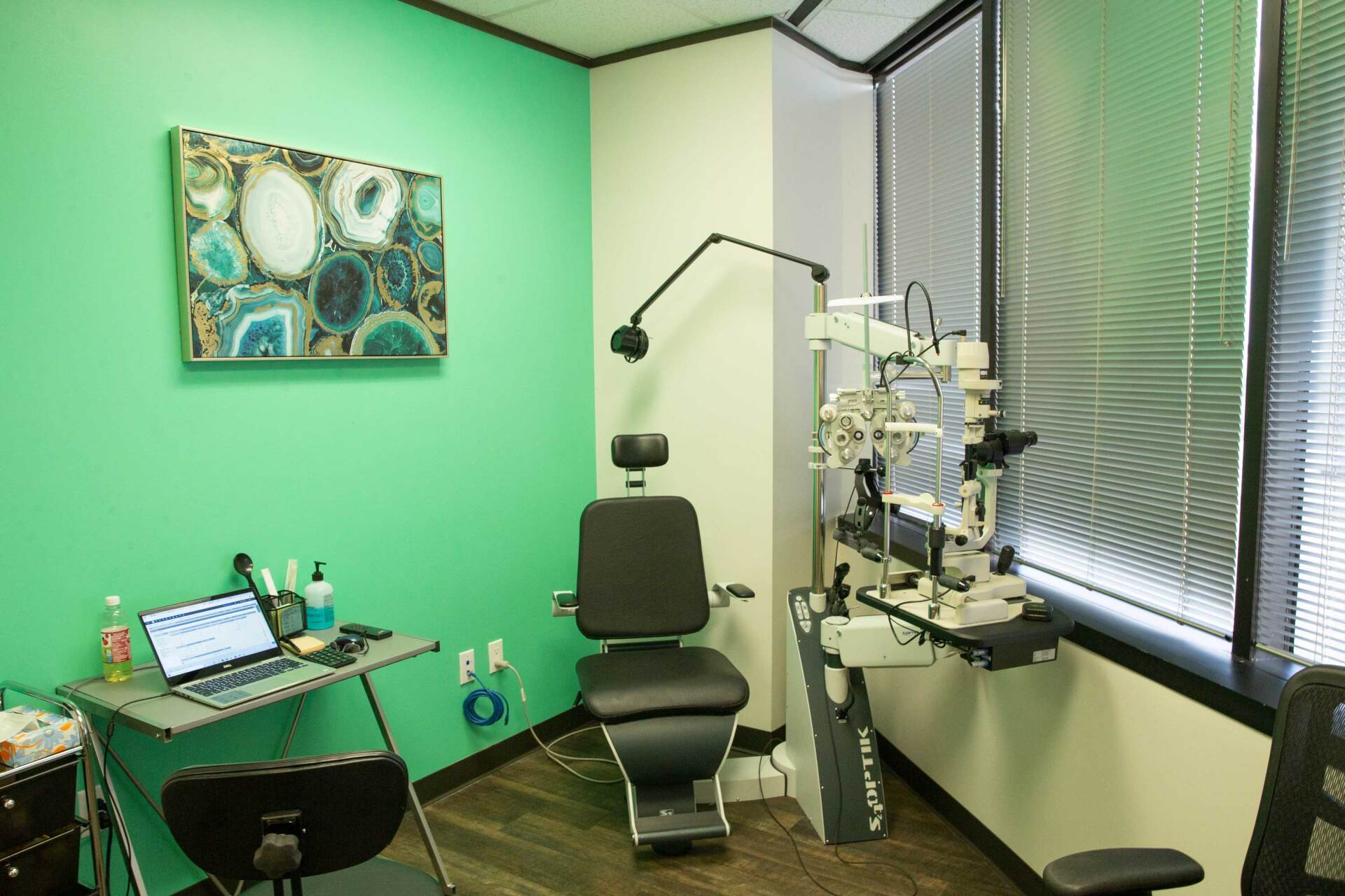 An eye exam room with a black exam chair, diagnostic equipment, a desk with a laptop, and a green accent wall with art.