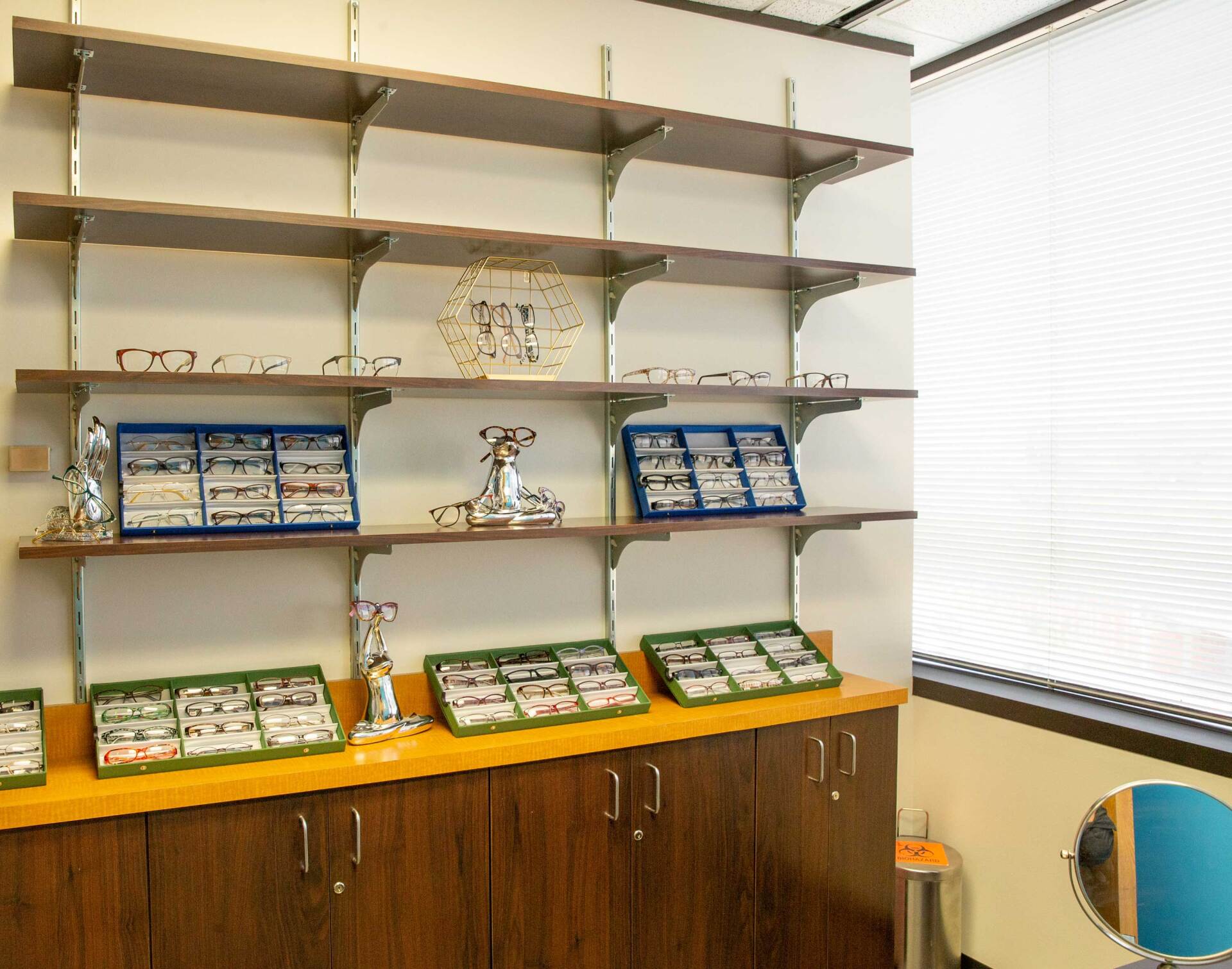 A wooden shelving unit and cabinets filled with various eyeglass frames in an office setting next to a window.