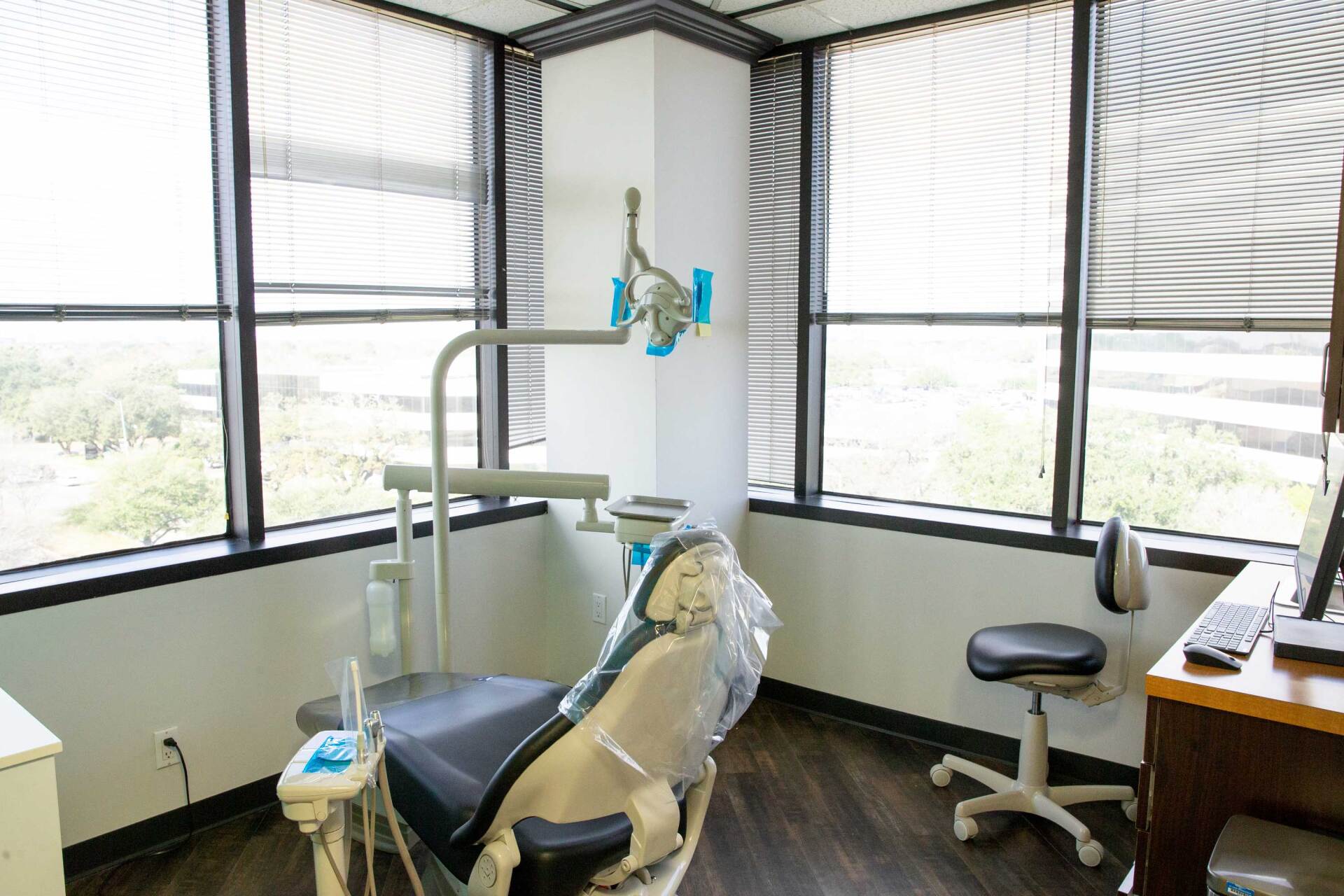 An empty dentist office with a patient chair, dental light, and a stool near a desk by large windows.