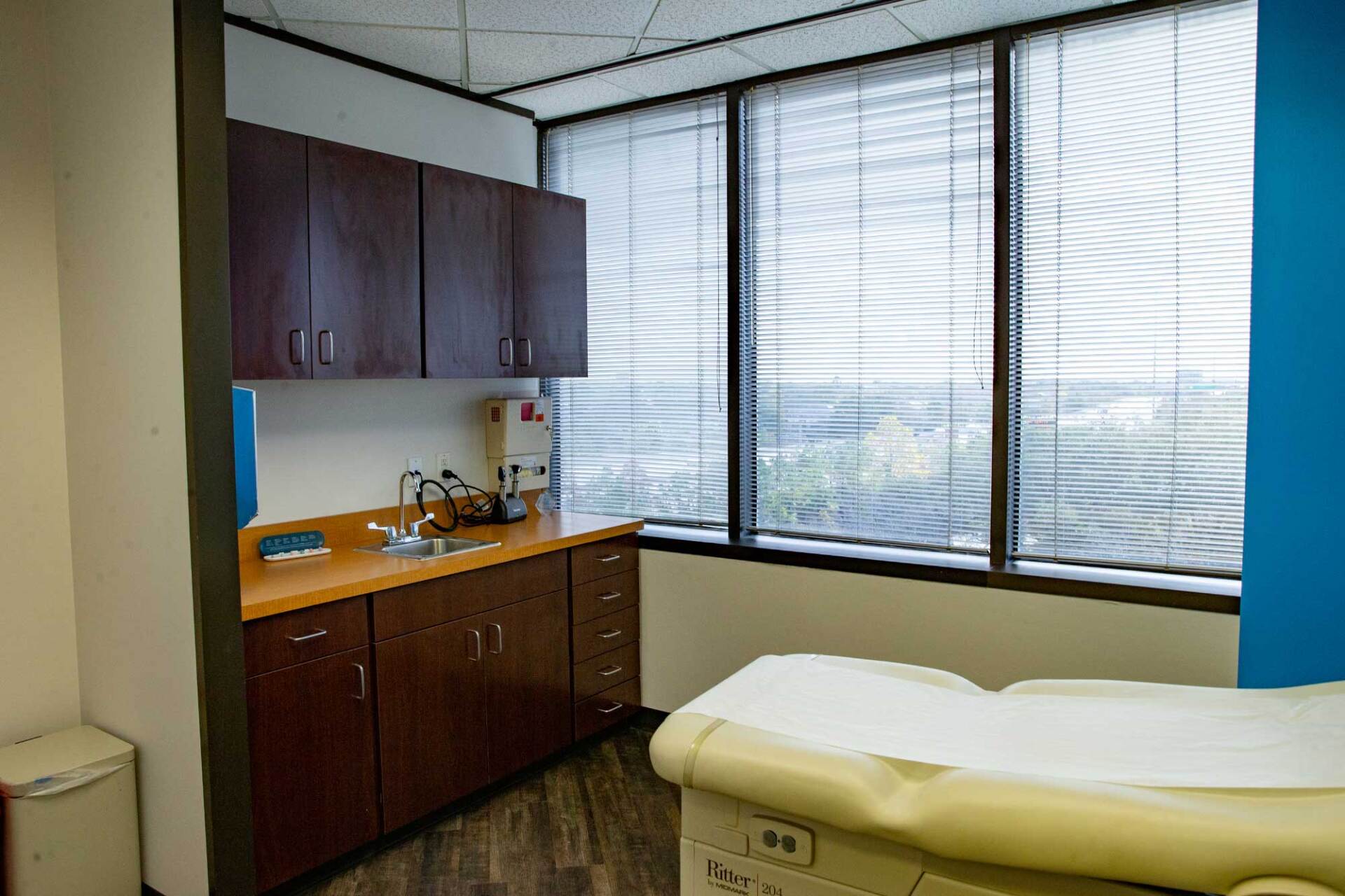 A medical examination room with dark wood cabinets, a sink, and a yellow padded exam table near a large window.