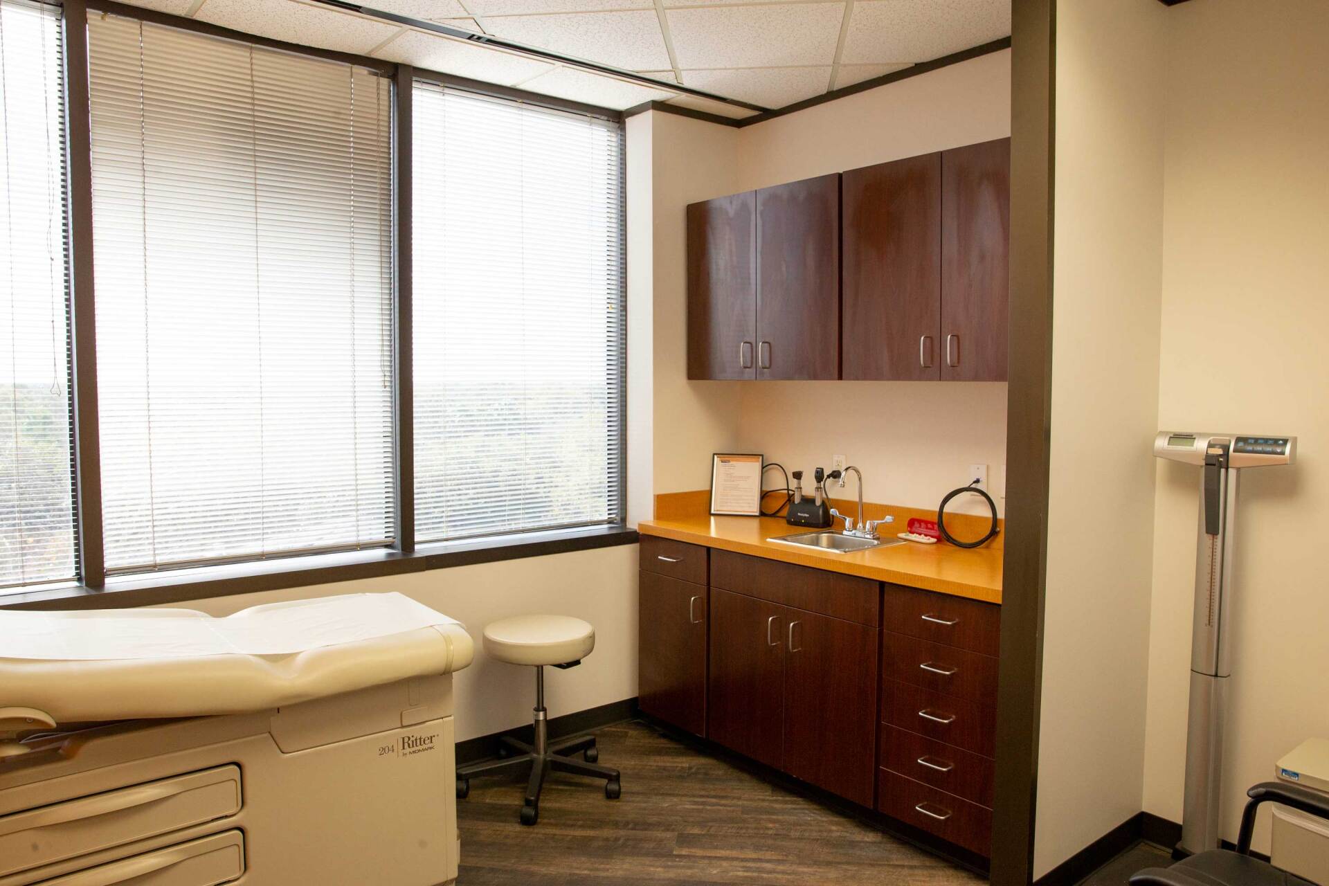 A medical examination room with an exam table, stools, dark wood cabinetry, a sink, and a medical scale near the window.