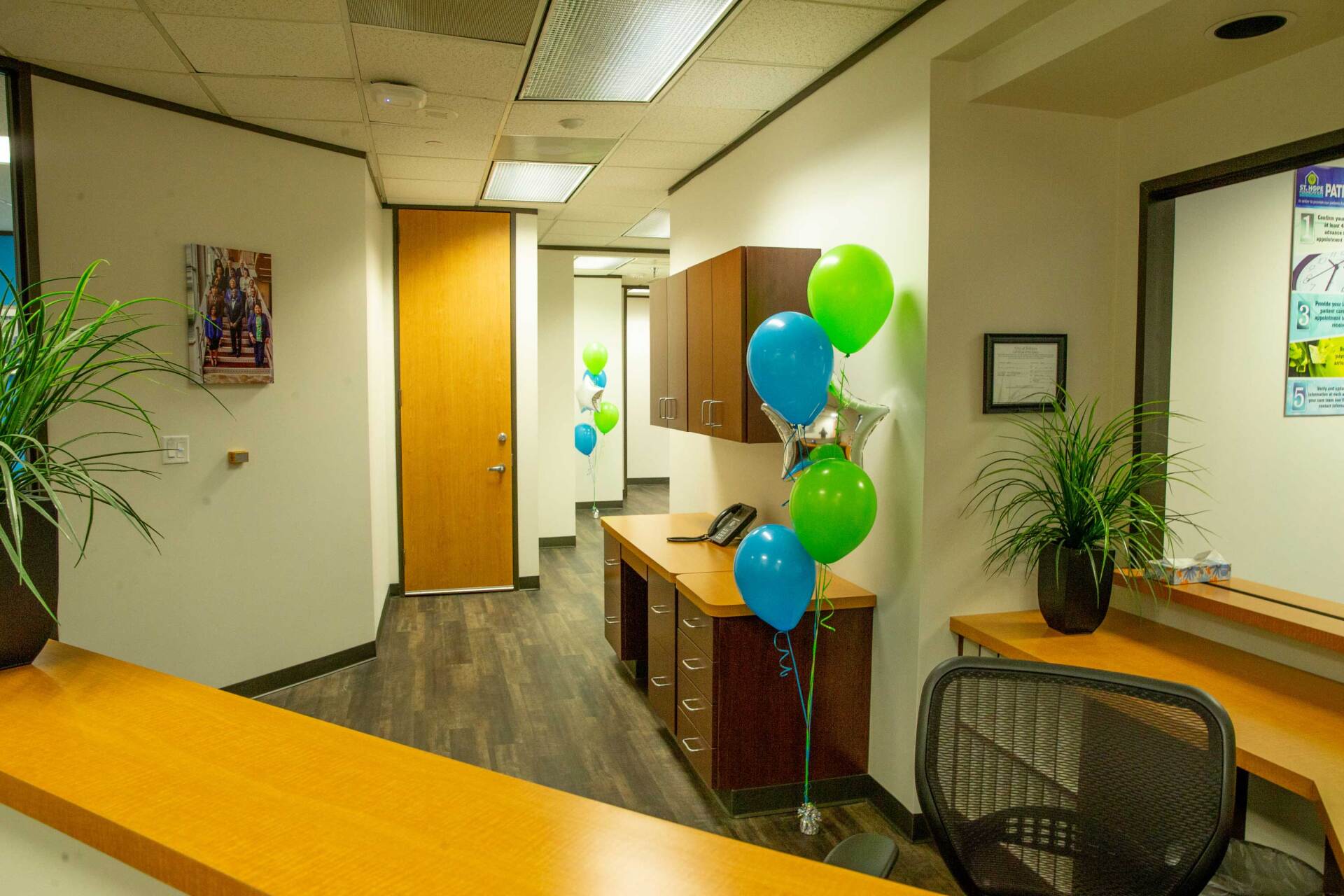 An office reception area with a wooden desk, a plant, and a bouquet of blue and green balloons.