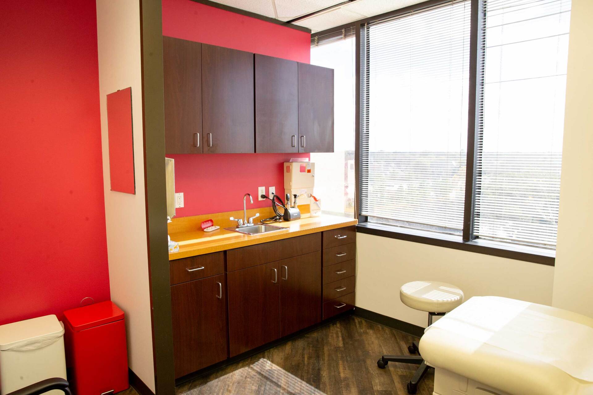 A medical exam room featuring dark cabinets, a sink, a white treatment table, a rolling stool, and bright red walls.