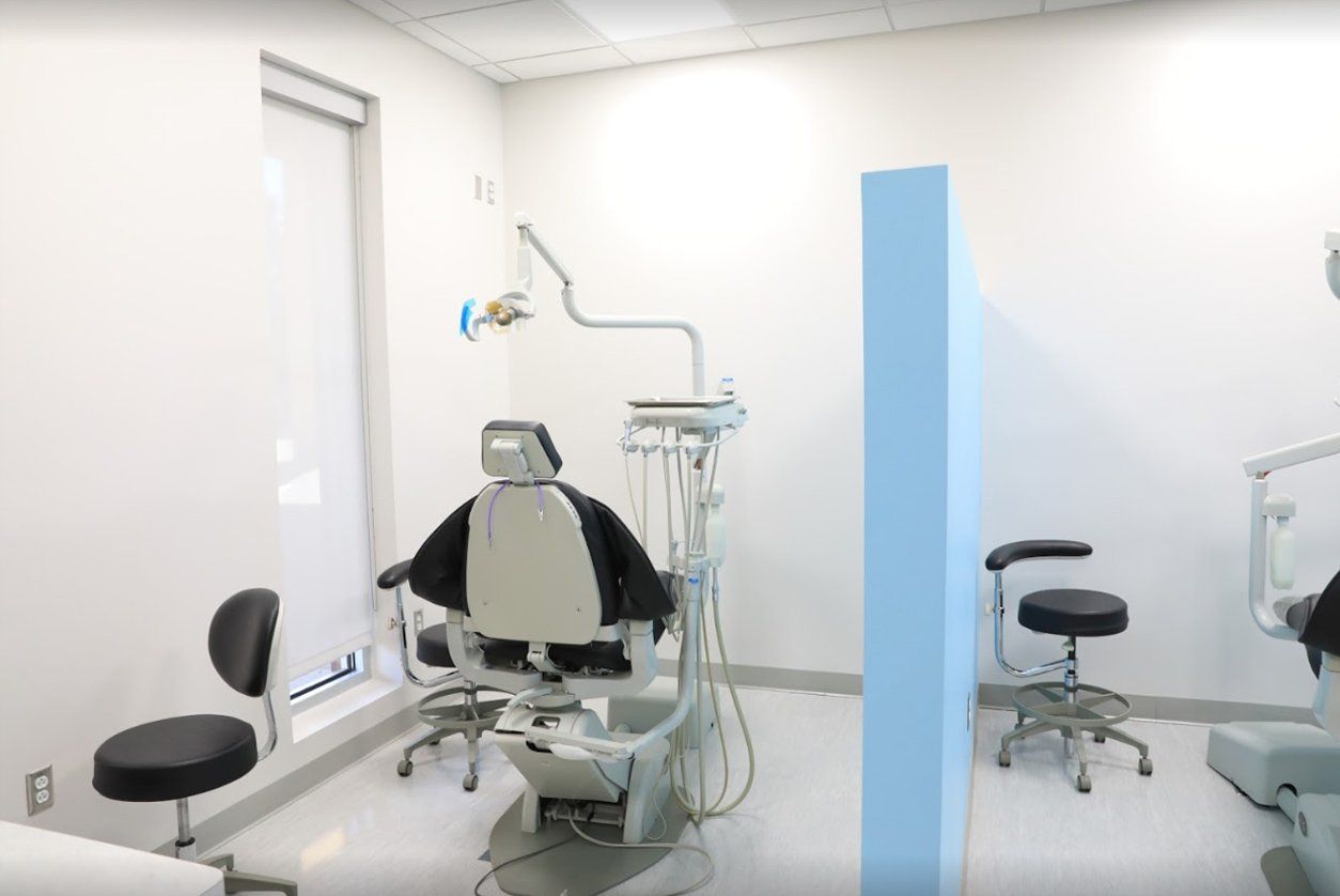 A bright dental exam room with two patient chairs, ergonomic stools, and overhead lighting separated by a blue partition.