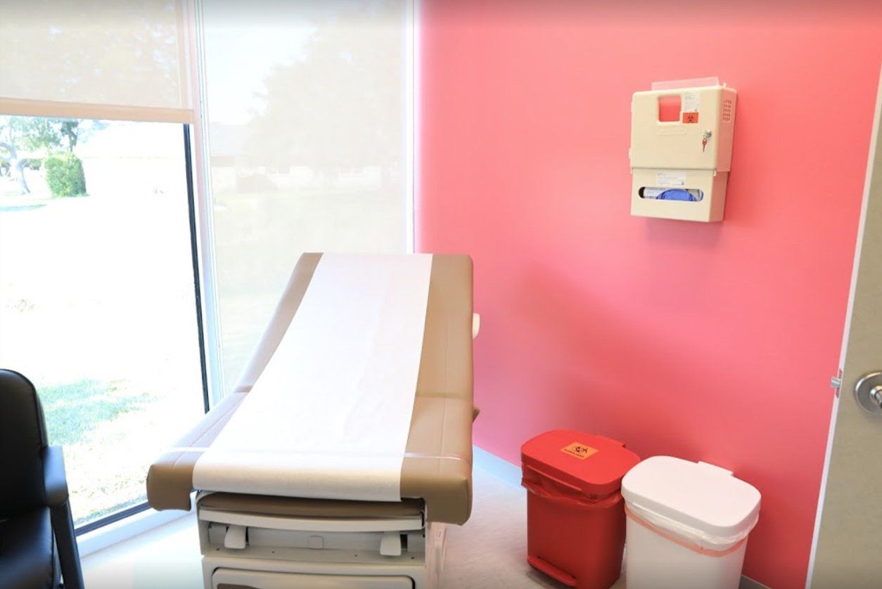 A medical exam room with a tan exam table, a window, a wall-mounted supply dispenser, and red and white waste bins.