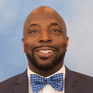 A professional headshot of a person wearing a dark blue suit, white shirt, and a blue polka-dot bow tie against a blue background.