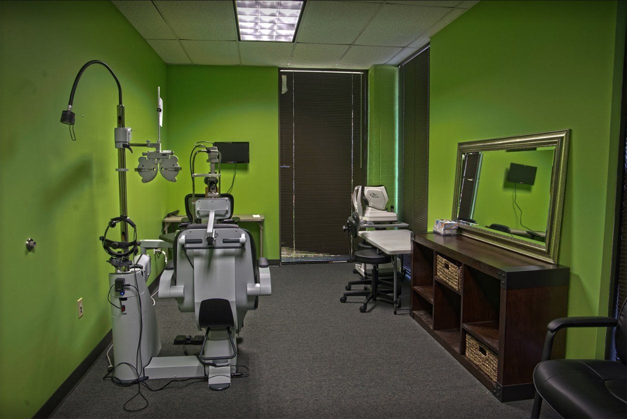 An optometry exam room with bright green walls, an examination chair, phoropter, and a mirror over a storage unit.