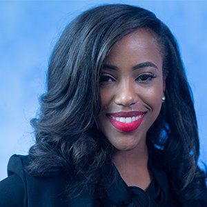 A professional headshot of a smiling person with dark, wavy hair, wearing a black blazer against a blue background.