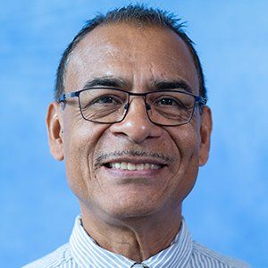 A smiling person with short dark hair, wearing rectangular glasses and a light-colored striped shirt against a blue background.