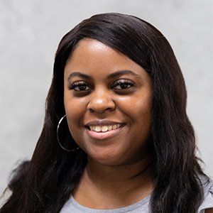A person with dark, wavy hair and hoop earrings smiling warmly against a light grey background.